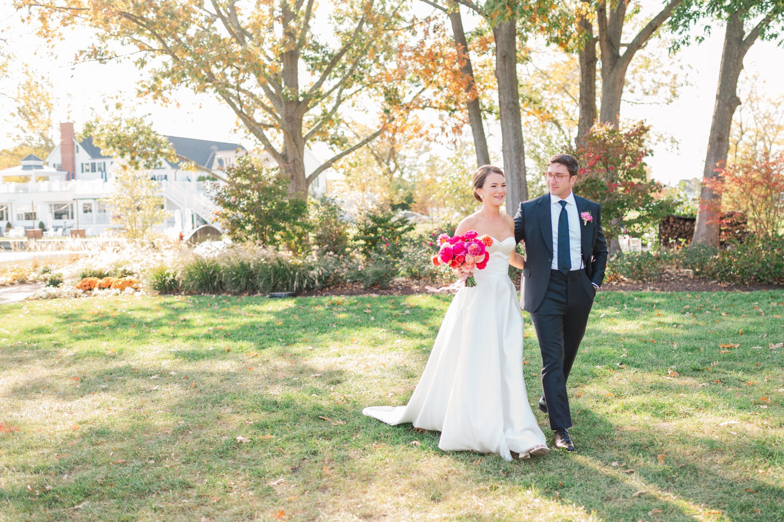 Bride and groom walk hand in hand on a sunny day at The Oaks Waterfront in Easton, Maryland. The bride wears a strapless silk wedding gown and carries a vibrant pink bouquet, while the groom looks at her lovingly in a classic black suit. The elegant waterfront venue with autumn foliage and a white estate is visible in the background.
